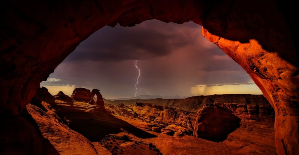 Uitzicht vanuit een grote rotsboog op een rood woestijnlandschap met Delicate Arch in de verte, terwijl een bliksemschicht inslaat onder een donkere onweerslucht.