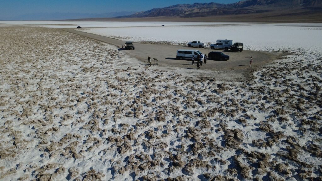 Uitzicht over Devil’s Golf Course in Death Valley National Park: een uitgestrekte zoutvlakte met ruige, puntige zoutformaties en diepe scheuren. Het oppervlak is onregelmatig en scherp, met witte, grijze en bruine tinten. Aan de horizon donkere woestijnbergen onder een heldere lucht.