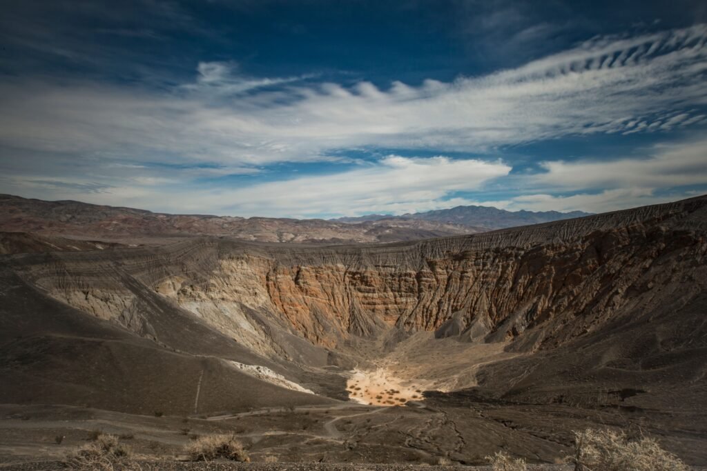 Uitzicht over Ubehebe Crater in Death Valley National Park: een enorme, komvormige vulkanische krater met steile wanden van donkere as, puimsteen en gelaagde rotslagen. De bodem ligt diep onder de rand. Rondom strekt zich droog woestijnlandschap uit met lage begroeiing en donkere bergen aan de horizon onder een heldere lucht.
